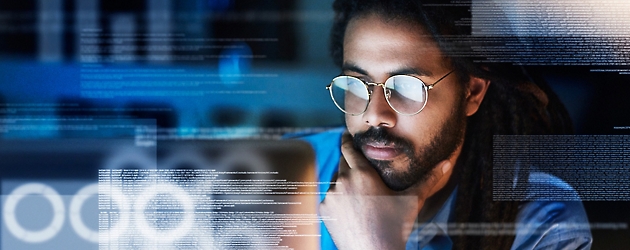 Man with long hair and glasses looking at computer screens displaying digital data overlays.