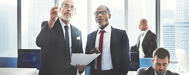 Two businessmen discussing over documents in a high-rise office, another man working in the background.