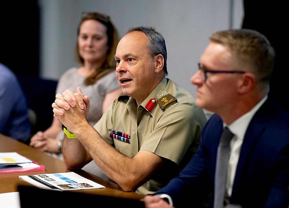 En mann i en militær uniform som snakker under et møte med to andre oppmerksomme deltakere i et konferanserom.