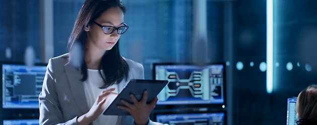 Una mujer con gafas revisa los datos de una tableta en una sala de control de tecnología apagada.