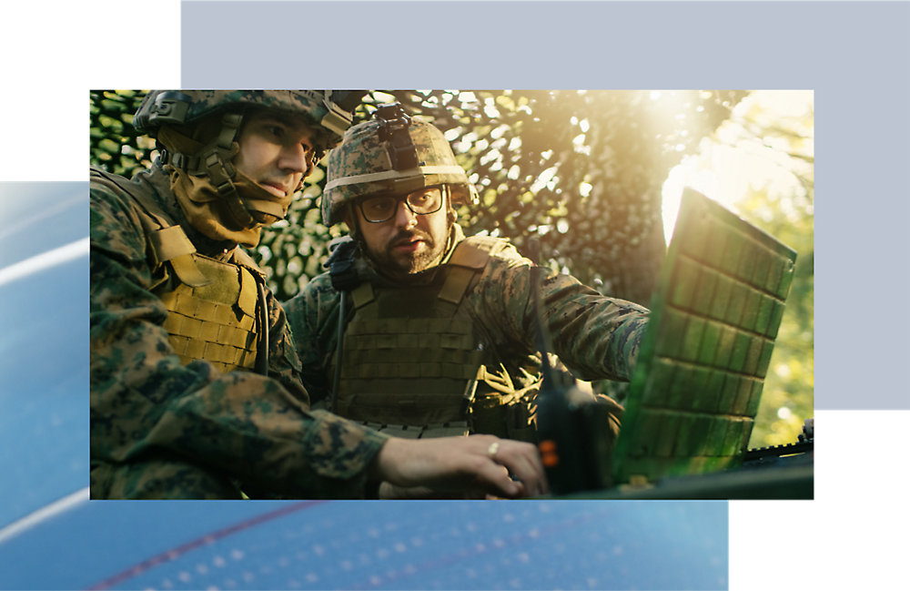 Dos soldados camuflados examinan un ordenador en un bosque, con la luz del sol filtrándose entre las hojas.