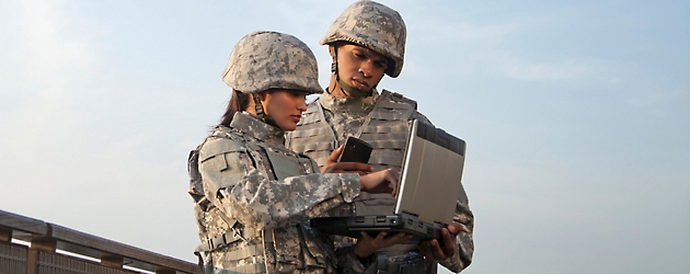 Zwei Militärangehörige, ein Mann und eine Frau, in Tarnuniformen benutzen ein tragbares Gerät im Freien.