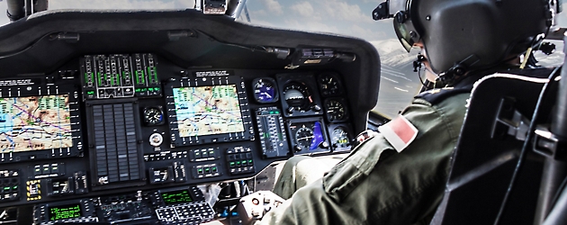 Пилот в кабине военного вертолета с подробными приборными панелями и несколькими экранами.