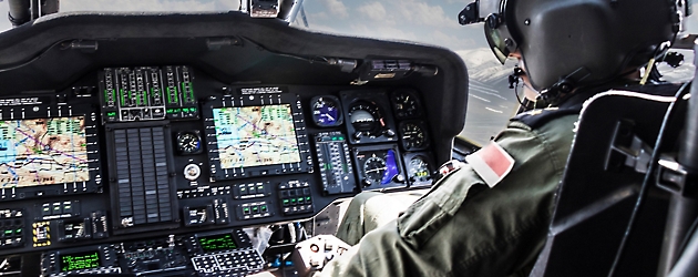 Pilot in military helicopter cockpit with detailed instrument panels and multiple display screens.