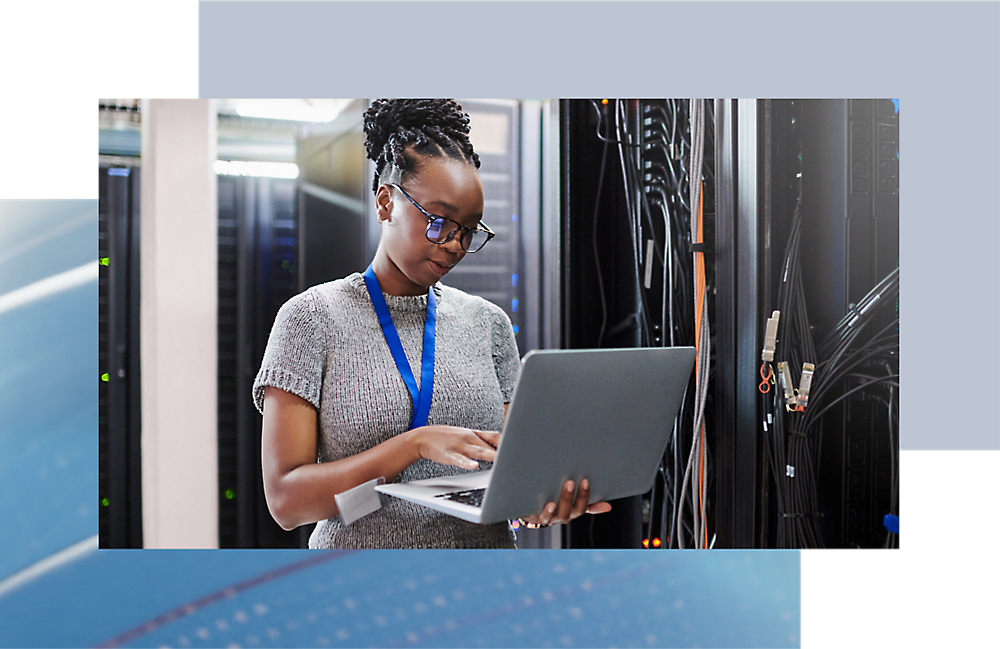 A woman in a server room analyzes data on her laptop.