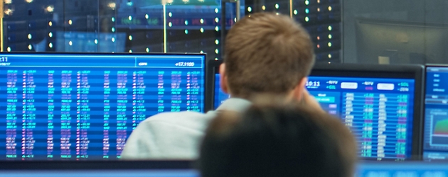 Rückenansicht von zwei Personen, die in einem Kontrollraum Finanzdaten auf mehreren blauen Bildschirmen überwachen.