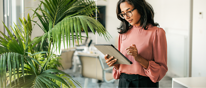 A professional woman in a pink blouse and glasses reading a tablet in a modern office with a large potted plant.