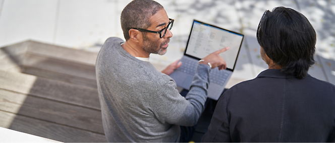 Two men sitting on outdoor stairs, one pointing at a laptop screen held by the other, in a discussion.