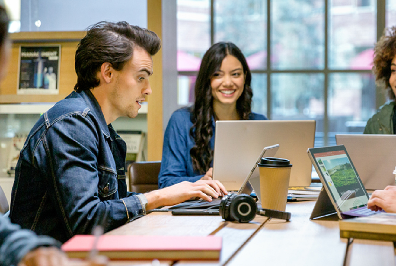 A group of four people sits at a wooden table with laptops, notebooks, and a coffee cup in front of them