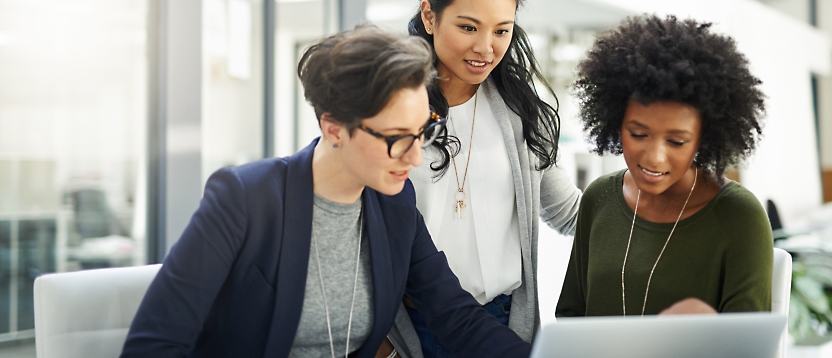 Three businesswomen of diverse ethnicities working together on a laptop in a modern office setting.