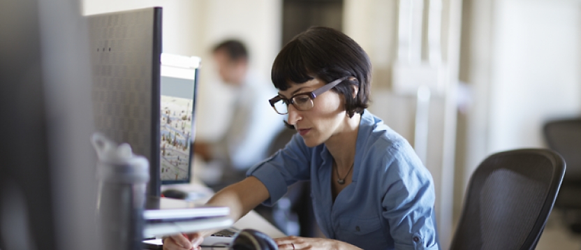 A woman with glasses, working intently at a computer in an office environment, with a colleague in the background.