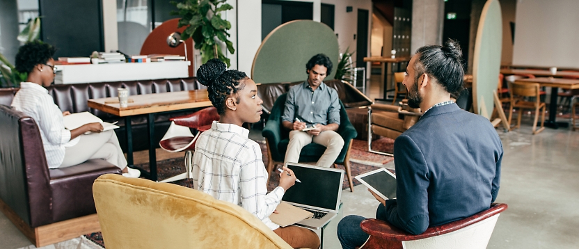 Four professionals in a casual meeting at a modern workspace, seated in chairs and sofas, engaging in a discussion.