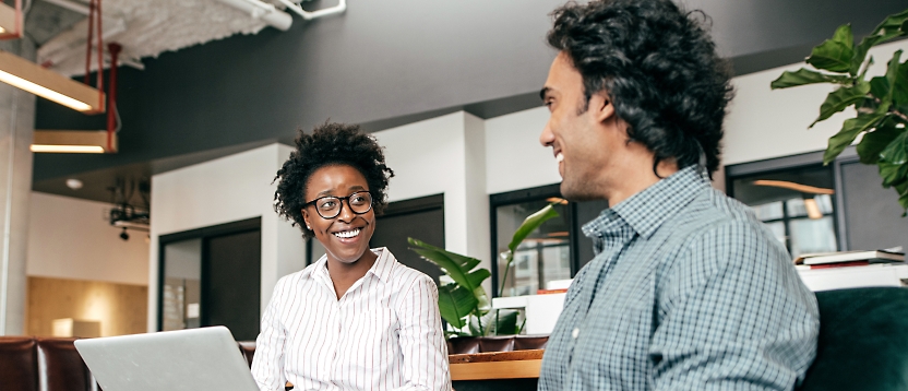 Two colleagues, a woman and a man, engaging in a cheerful conversation at a workspace with a laptop open