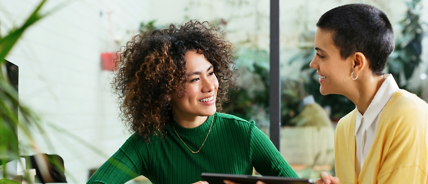 Two women engaged in a cheerful conversation in a plant-filled room, one holding a digital tablet.