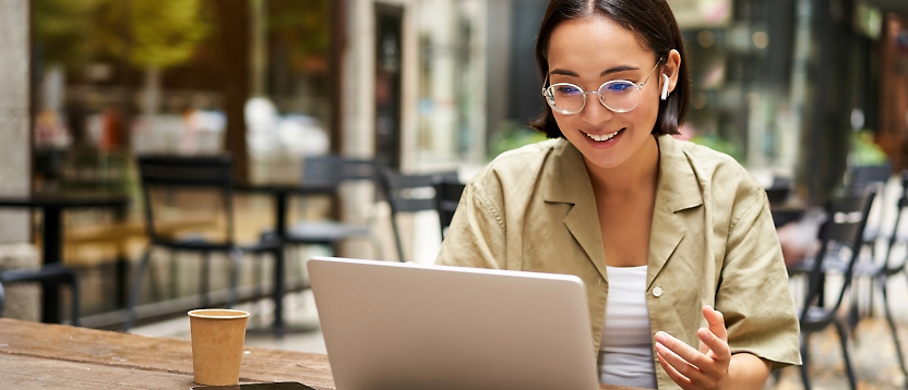 Young woman in glasses smiling and working on a laptop at a cafe table, with a disposable coffee cup beside her.