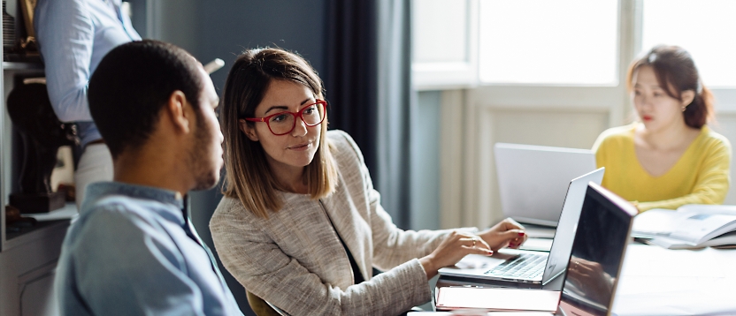 Two professionals, a man and a woman, engaged in a discussion with a laptop in between them in an office setting.