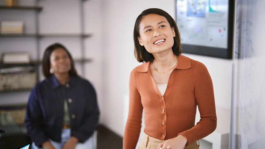 Deux femmes dans un bureau, une debout au premier plan souriant vers la caméra et une autre assise en arrière-plan.