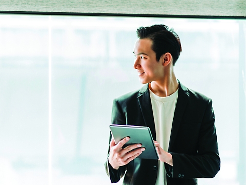 A person in a suit holding a tablet and looking to the side in a brightly lit room with large windows in the background.