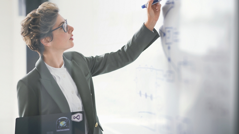 Eine berufstätige Frau mit kurzen Haaren in einem Blazer, die auf einem Whiteboard in einem hell beleuchteten Büro schreibt.