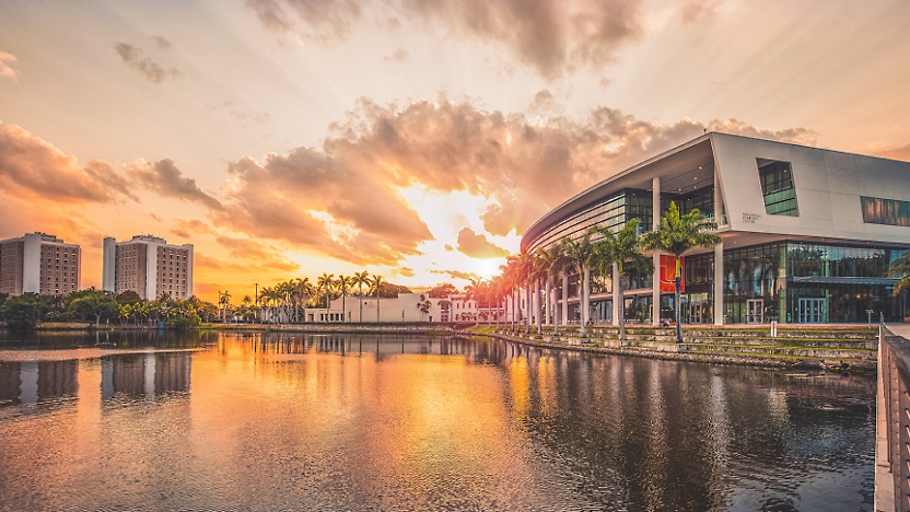 Sunset reflecting off a calm lake with modern buildings on the shore, under a vibrant sky with scattered clouds.