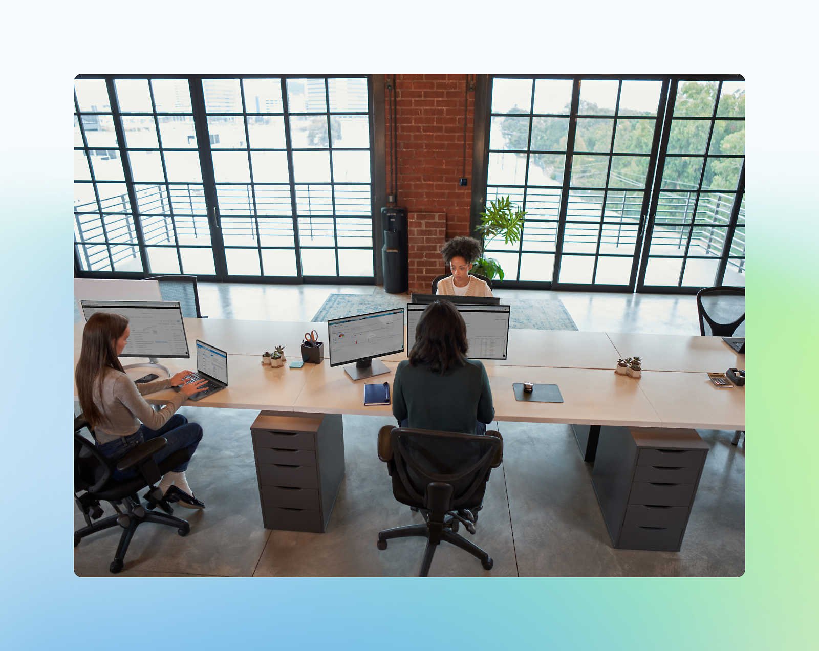 Three people work at computers in a modern office with large windows and a brick wall.