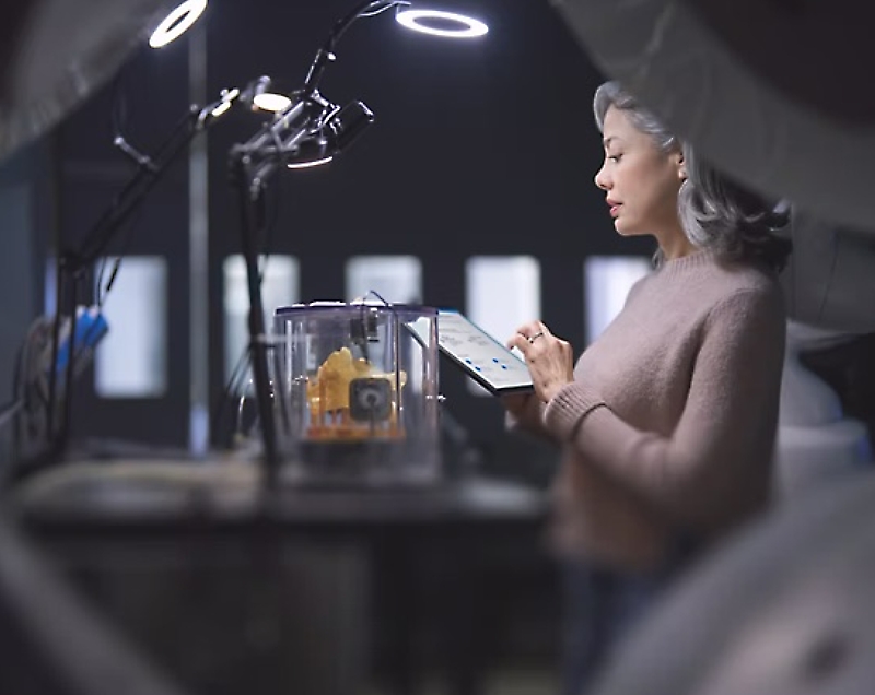 A woman examining a 3d print while holding a tablet in a workshop setting.