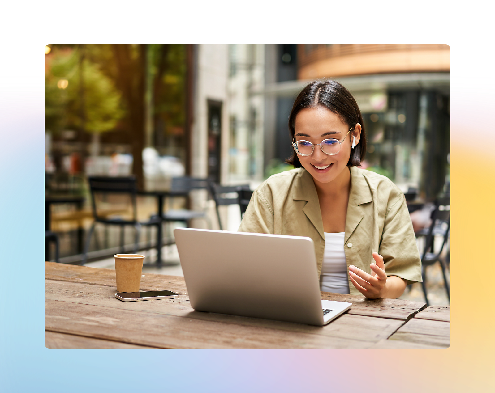 Person wearing glasses and a beige shirt, sitting at an outdoor table, smiling while using a laptop