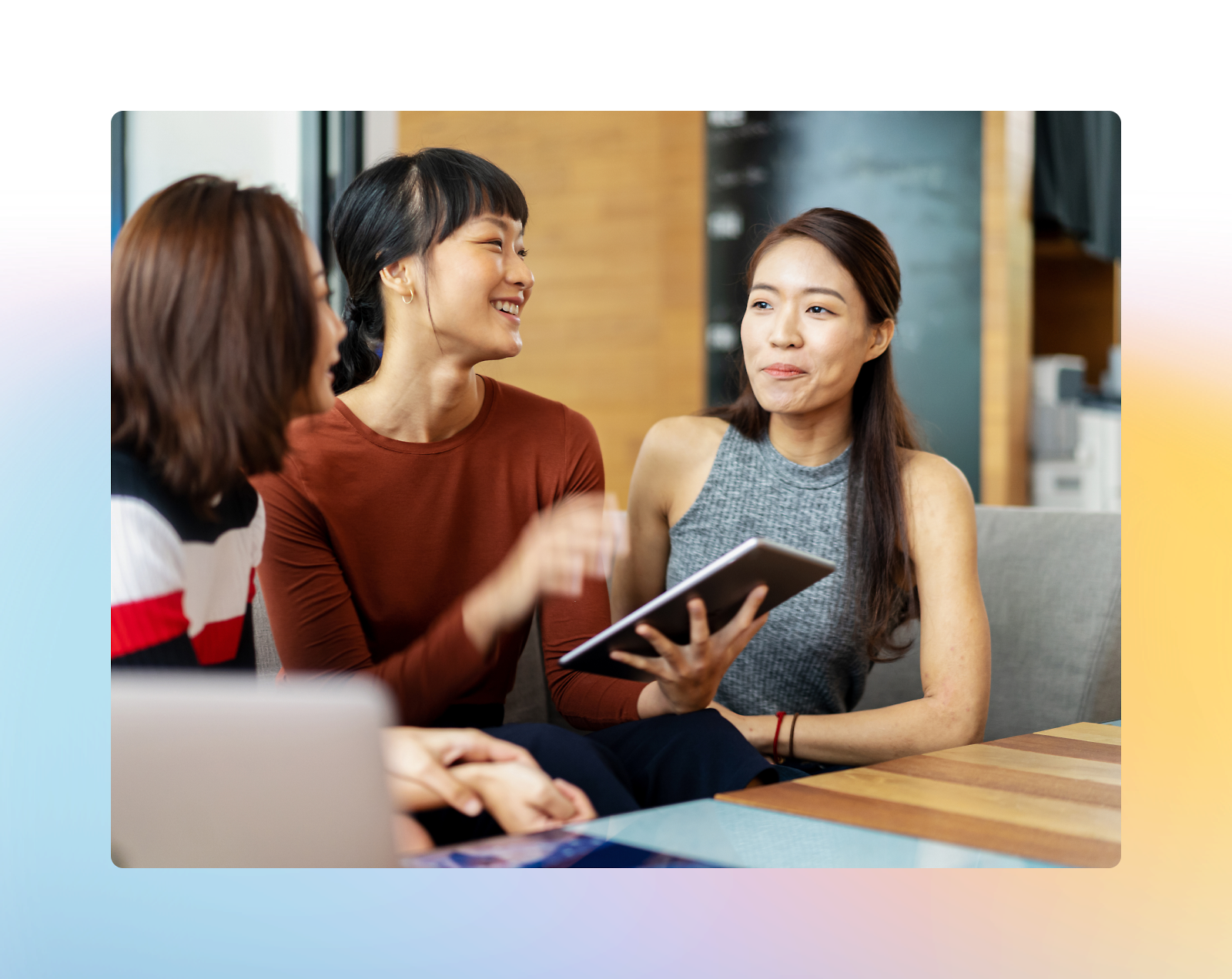 Three women are sitting together and engaging in a conversation. One woman holds a tablet while the others listen and smile.