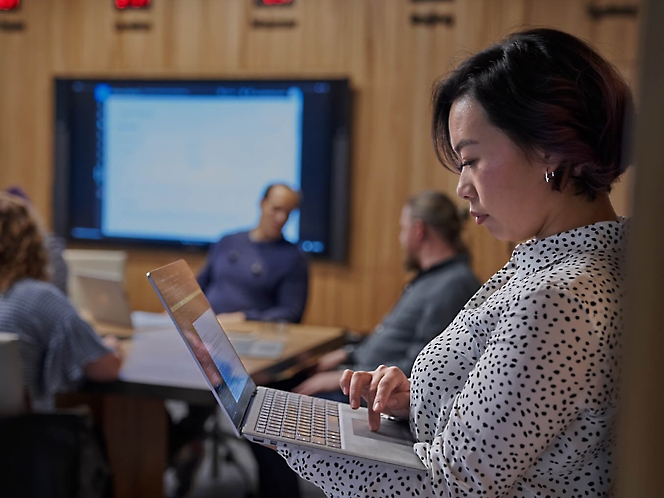 A professional woman working on a laptop in a conference room with colleagues in the background..