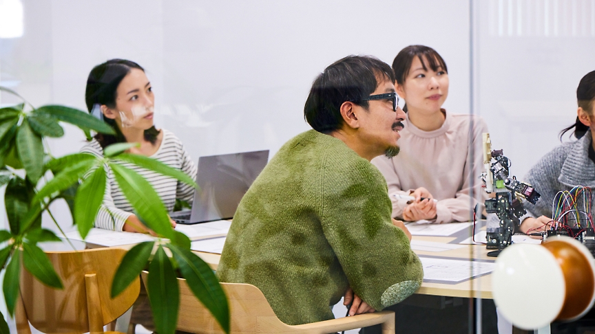 Four people sitting around a table in a meeting room, engaged in discussion. A laptop and documents are on the table.