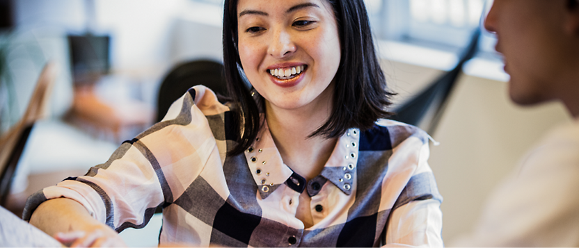 A woman with shoulder-length dark hair wearing a checkered blouse is smiling and interacting with another person 