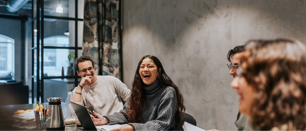 Three people enjoying a conversation at a table in a cafe, one woman laughing joyfully with a laptop open in front of them.