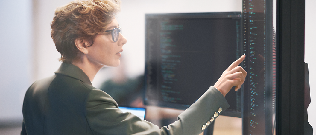 A woman with glasses analyzes code on multiple computer screens in a modern office setting.