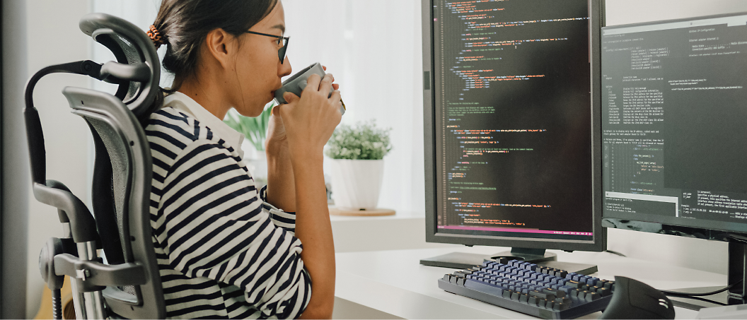 A woman in a wheelchair drinking coffee while looking at multiple computer screens displaying code.