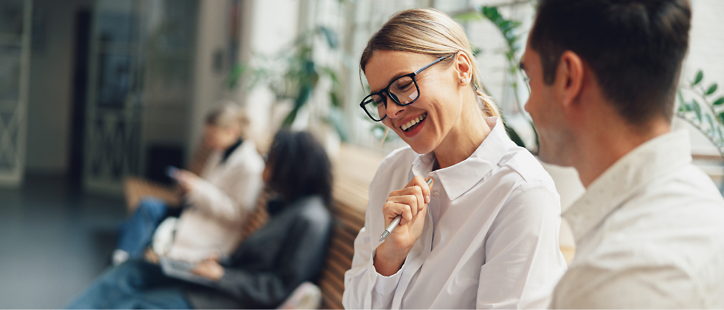 A woman in glasses smiles and talks with a man in a light office setting, both dressed in white shirts