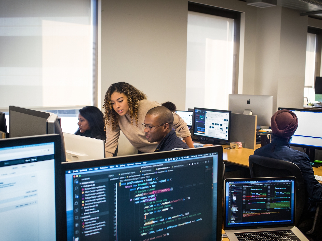 Группа разнообразных специалистов работает за компьютерами в офисной обстановке, глядя на экраны, на которых отображается различный программный код и данные.