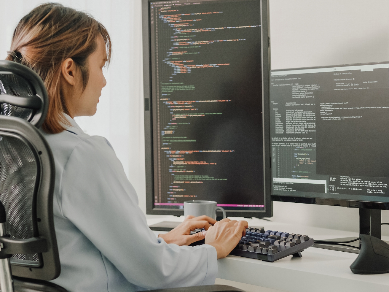 Een vrouw die op een bureaustoel zit en codeert op een computer met meerdere schermen met programmeercode.