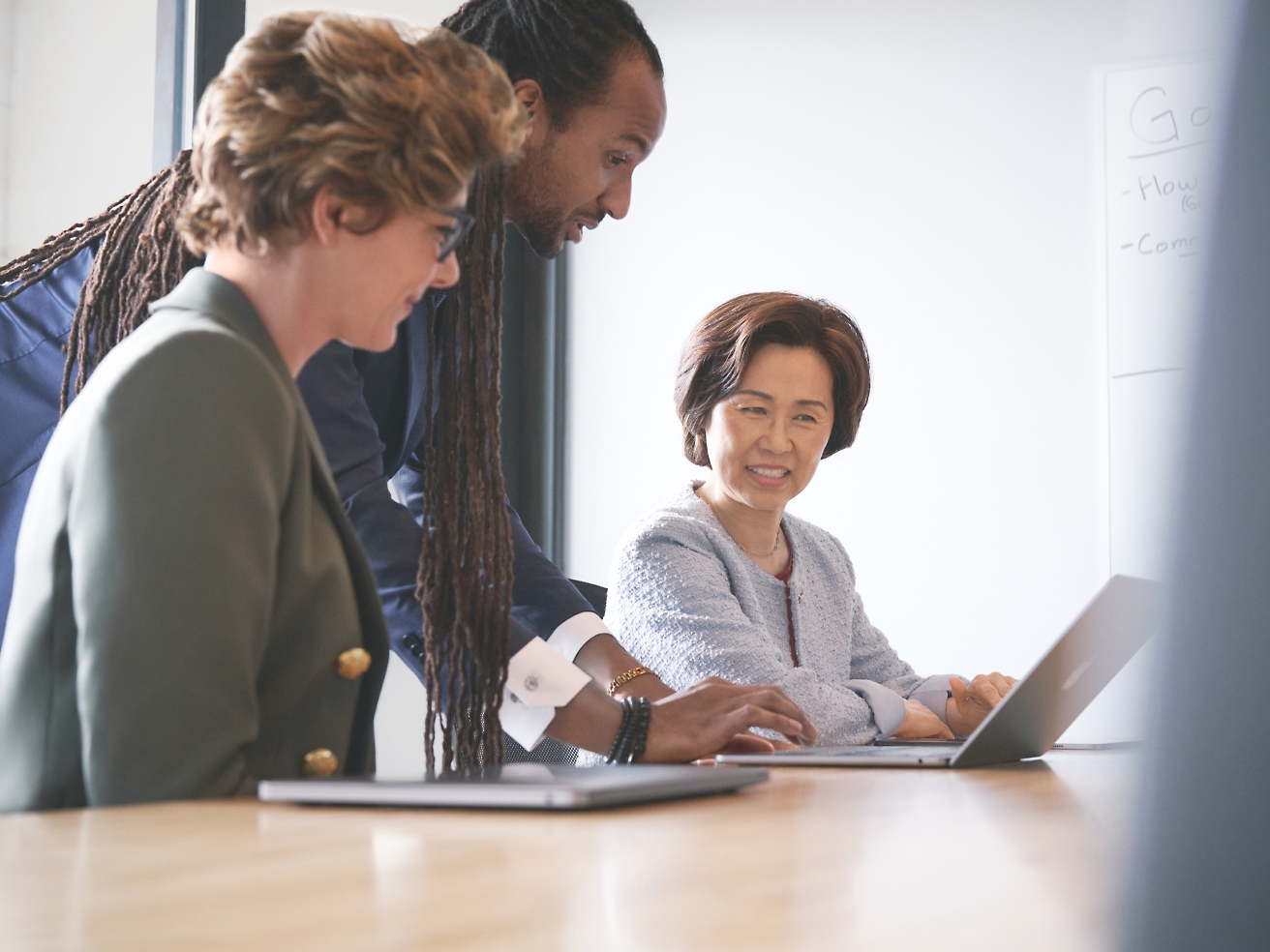 Three professionals, one with dreadlocks, sitting around a laptop in a meeting room, discussing work contentedly.