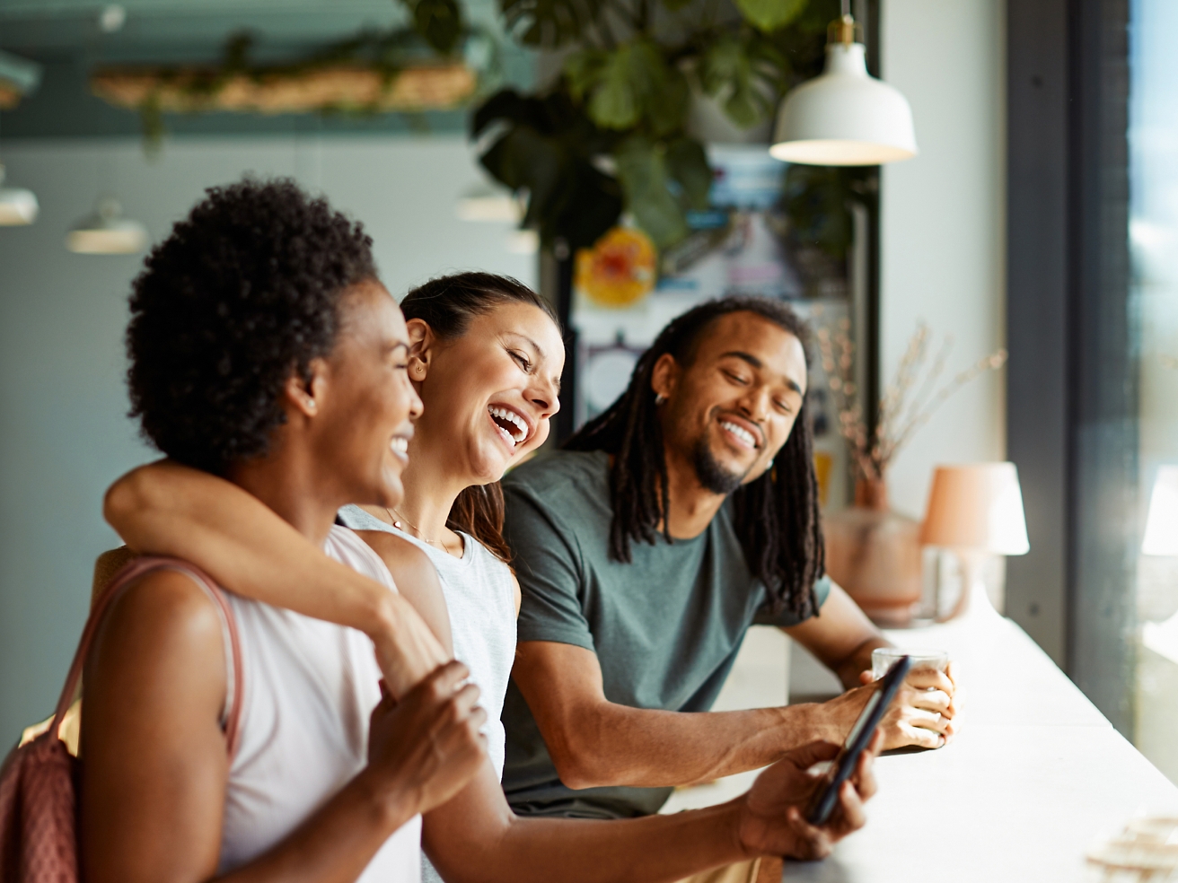 Trois amis riant et regardant un smartphone ensemble dans un café moderne.