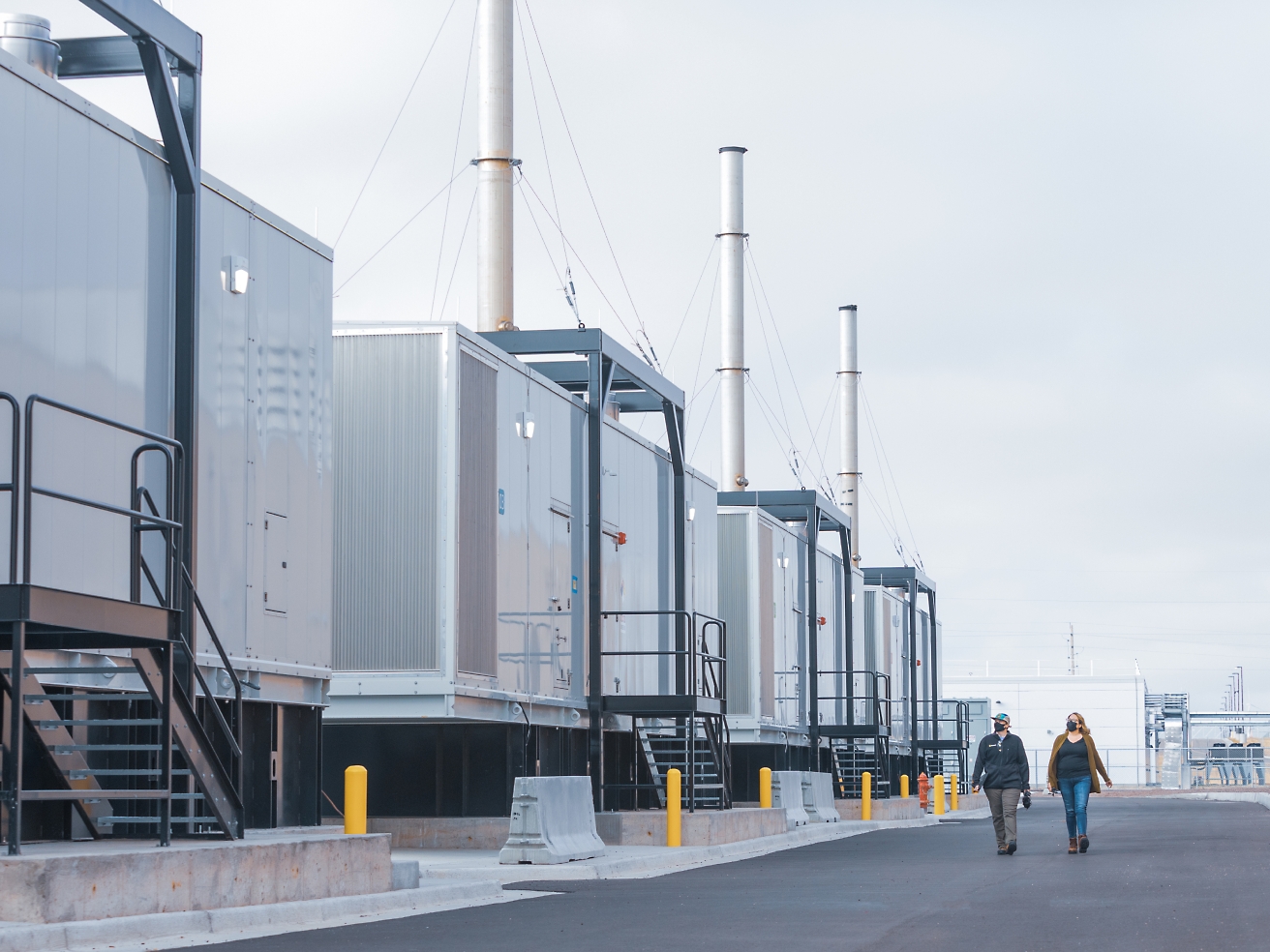 Twee personen die langs een rij industriële containers lopen, er zijn hoge schoorstenen van een installatie te zien en de lucht is bewolkt.