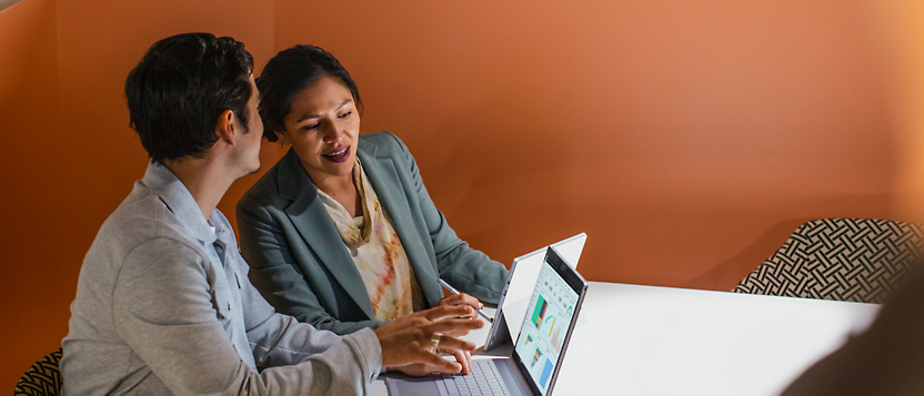 Two people sitting at a table, smiling and looking at a laptop screen together in a room with orange walls.