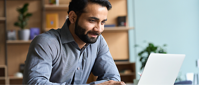 A man is sitting at a desk, looking at a laptop screen, and smiling.