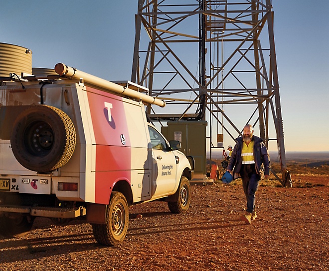 A utility worker walks towards a telecommunications tower carrying equipment