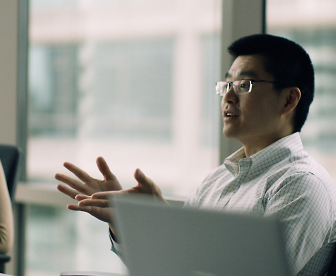 Person wearing glasses and a white checkered shirt speaks while gesturing with their hands in an office setting