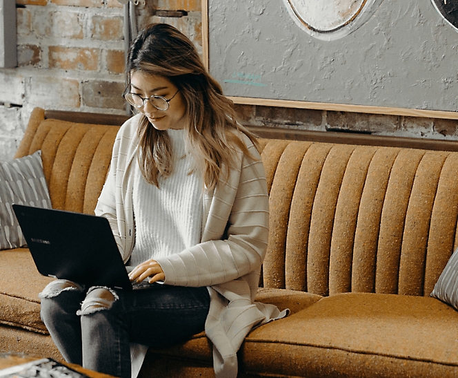 Женщина в очках и полосатом свитере сидит на горчичном диване и работает за ноутбуком в уютном кафе.