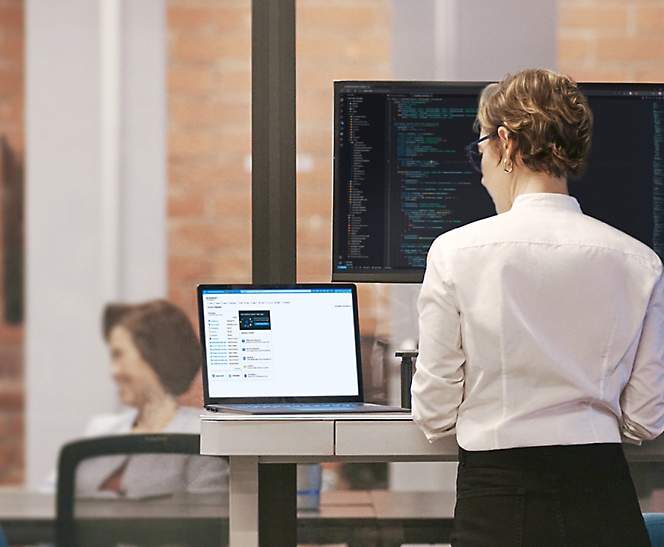 A woman stands at a standing desk, working on a laptop with a large monitor displaying code in the background.