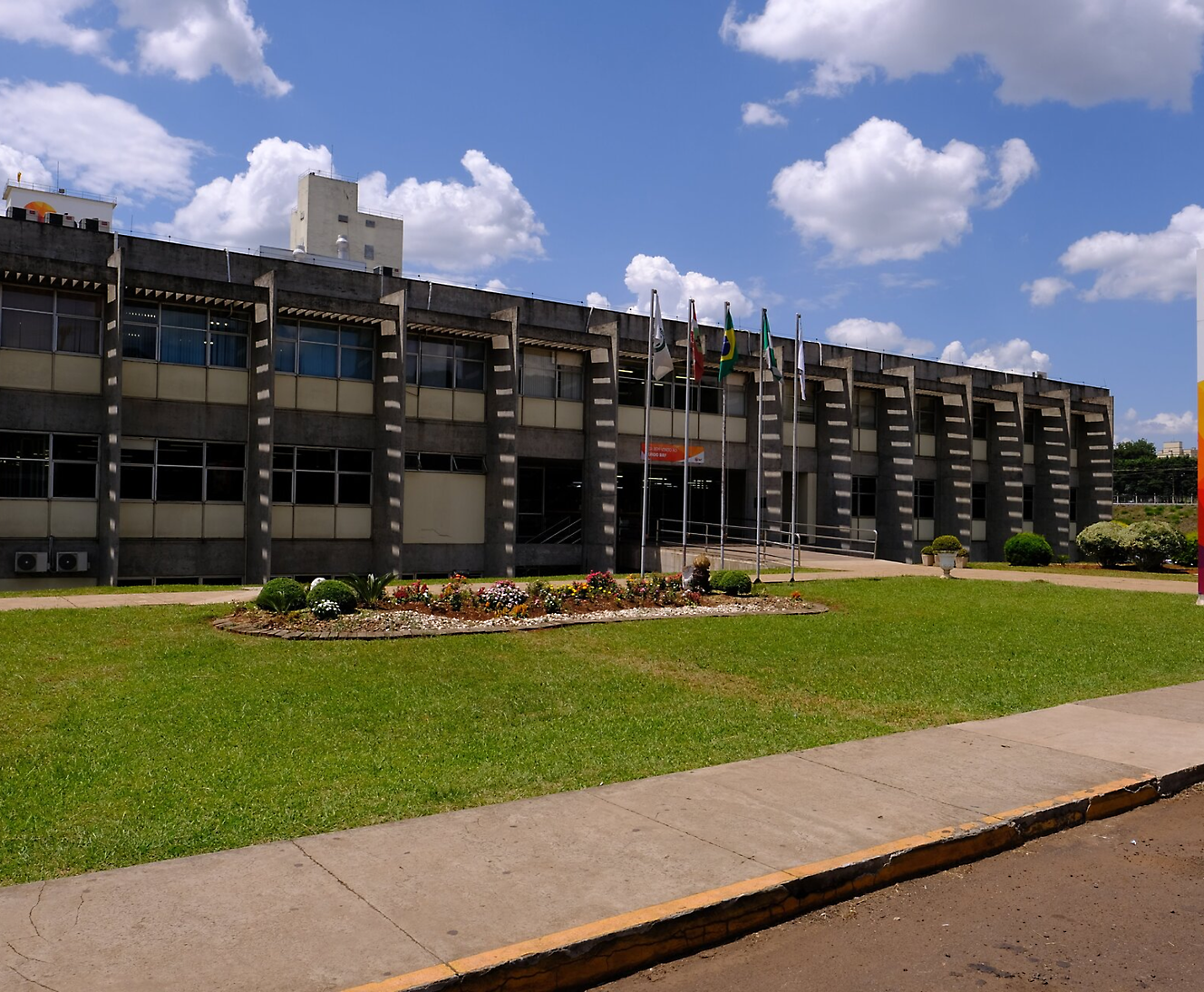 A modern, rectangular building with multiple windows and a landscaped garden in front, featuring flags on tall pole