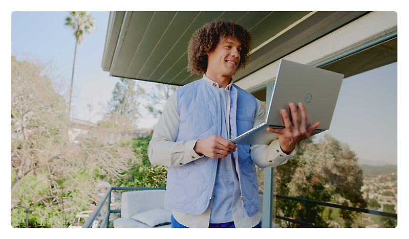 A man with curly hair using a laptop and holding papers stands on a balcony with greenery in the background.