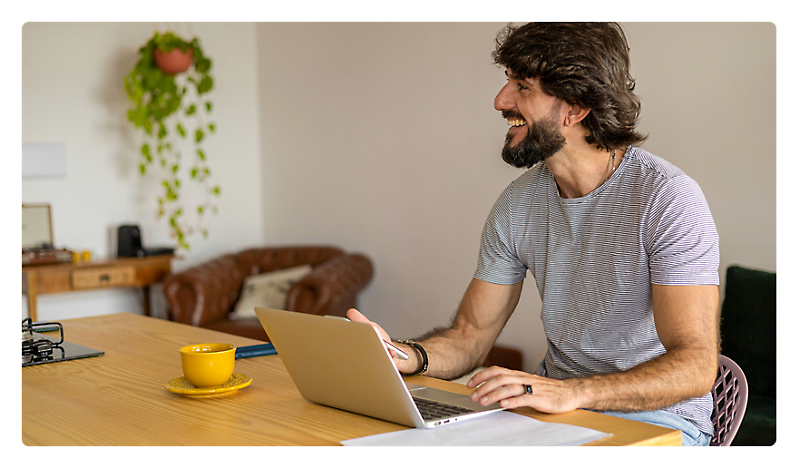 A man with a beard smiling while using a laptop at a home office desk with a cup of coffee and plant decor.