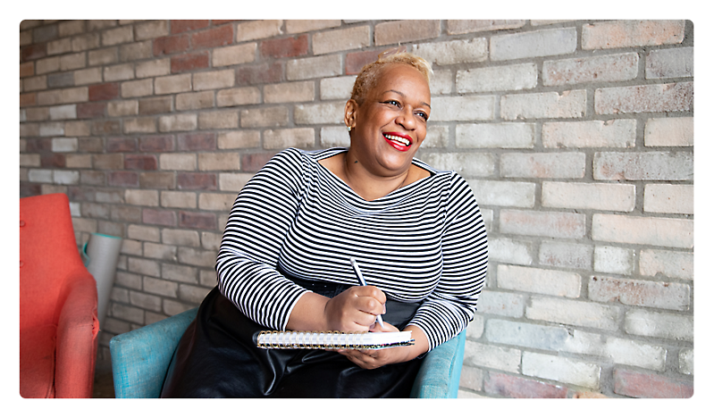 A joyful woman with short blonde hair, wearing a striped top, sitting in a teal armchair, writing in a notebook